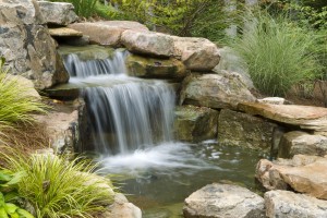 Long exposure of a backyard waterfall and pond surrounded by green foliage.